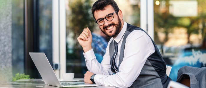 Young attractive successful caucasian elegant bearded businessman in suit with eyeglasses sitting in cafe, leaning on table and looking at camera.